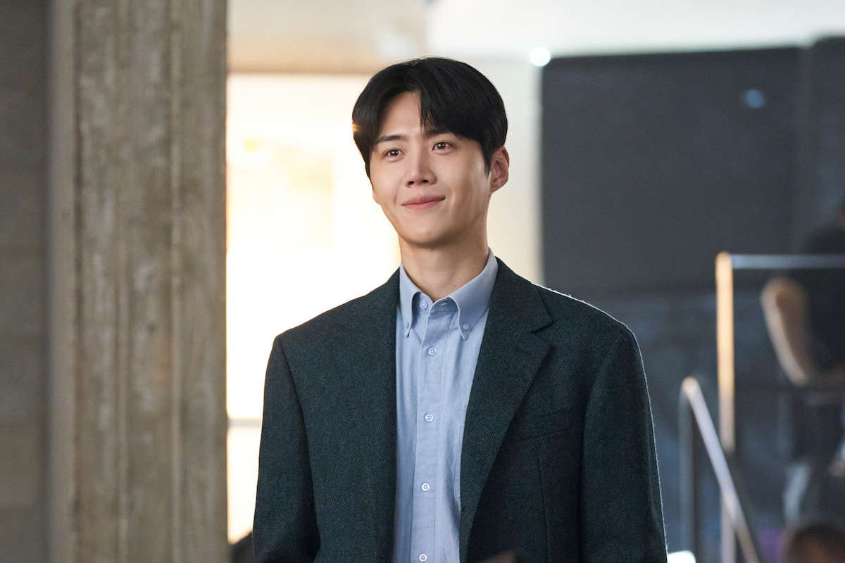 A young man in a suit stands indoors, smiling softly in a modern, warmly lit setting with a concrete pillar and blurred backgrounds, creating a calm and approachable mood.