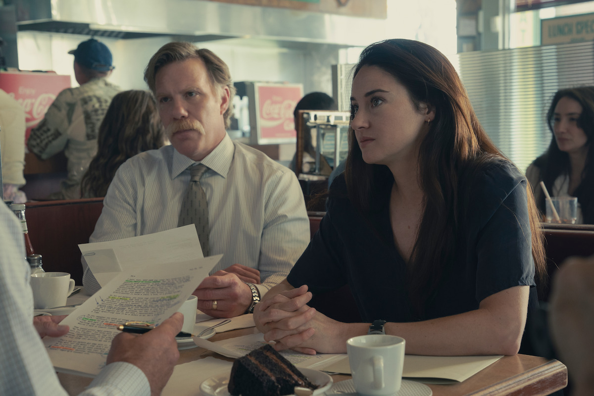 Two people sit at a diner table with coffee and cake, attentively listening to a third person who is holding documents. The setting is casual with retro decor, other patrons are visible in the background.