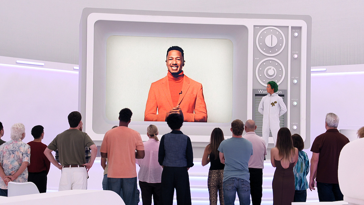 A group of people in a brightly lit modern room stand facing a giant retro TV displaying a man in an orange suit, while a person in a white lab coat stands beside the screen.