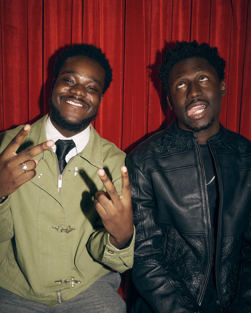 Two men posing in front of a red curtain; one is smiling and making peace signs with his hands, while the other rolls his eyes and wears a black jacket. Indoor setting, casual and playful atmosphere.