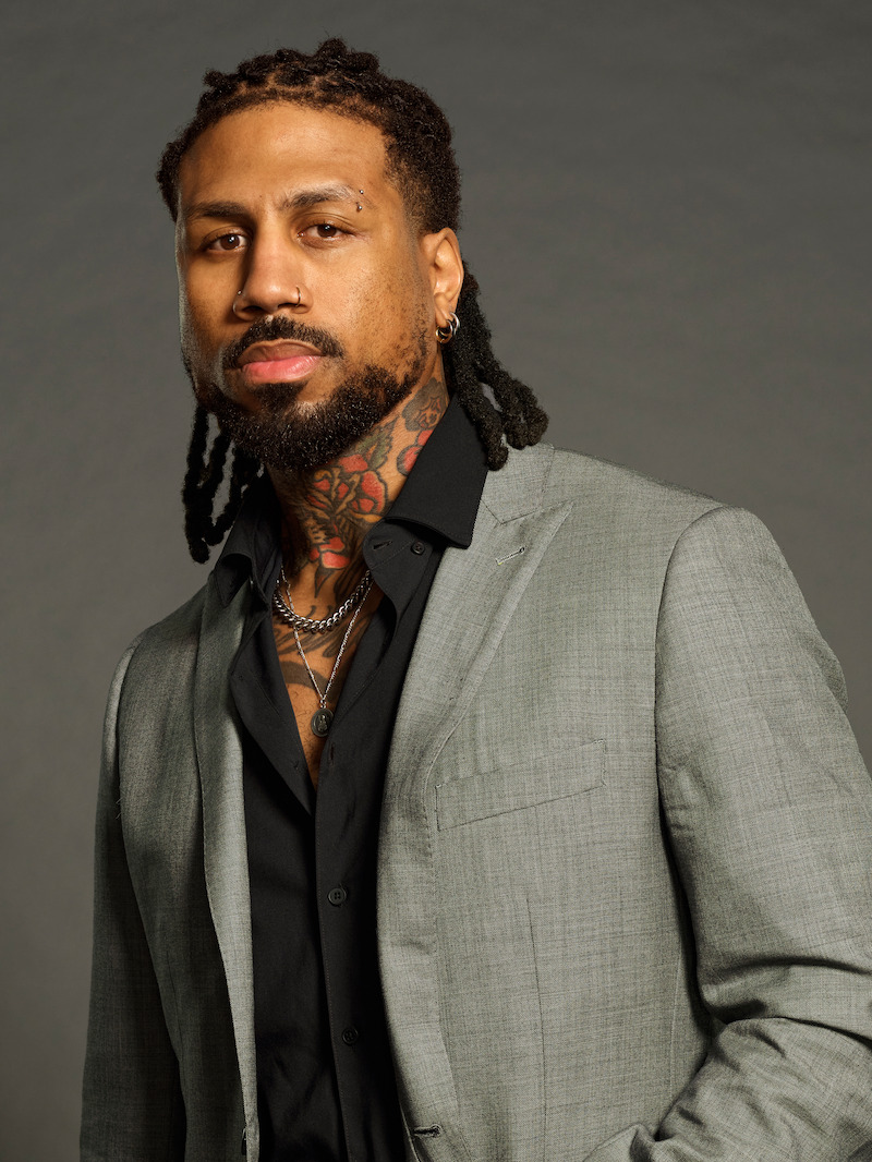 Man with dreadlocks, beard, and neck tattoos wearing a light gray suit and black shirt, layered necklaces, standing in front of a plain, neutral background with soft lighting, posing confidently.