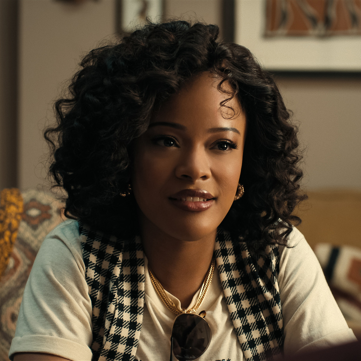Woman with curly hair sitting indoors, wearing a checkered vest over a white shirt, gold jewelry, and sunglasses hanging from her neck. Cozy living room setting with patterned cushions and framed art on the wall in the background.