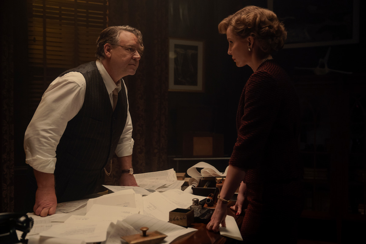 A man and woman in formal attire have a serious discussion in a dimly lit office, standing over a desk covered with papers and old-fashioned equipment, creating a tense atmosphere.