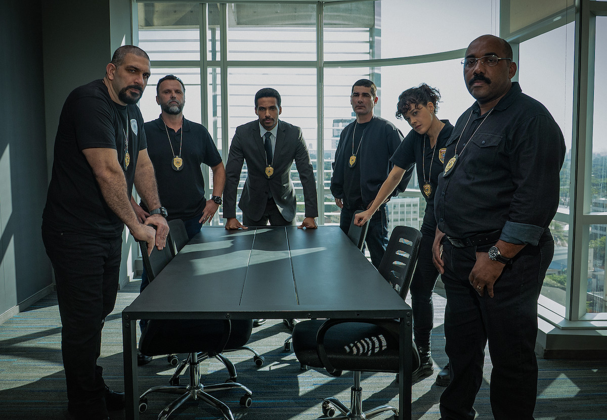 Grupo de 5 homens e 1 mulher, com distintivos no pescoço, se reúne em volta de uma mesa de delegacia policial