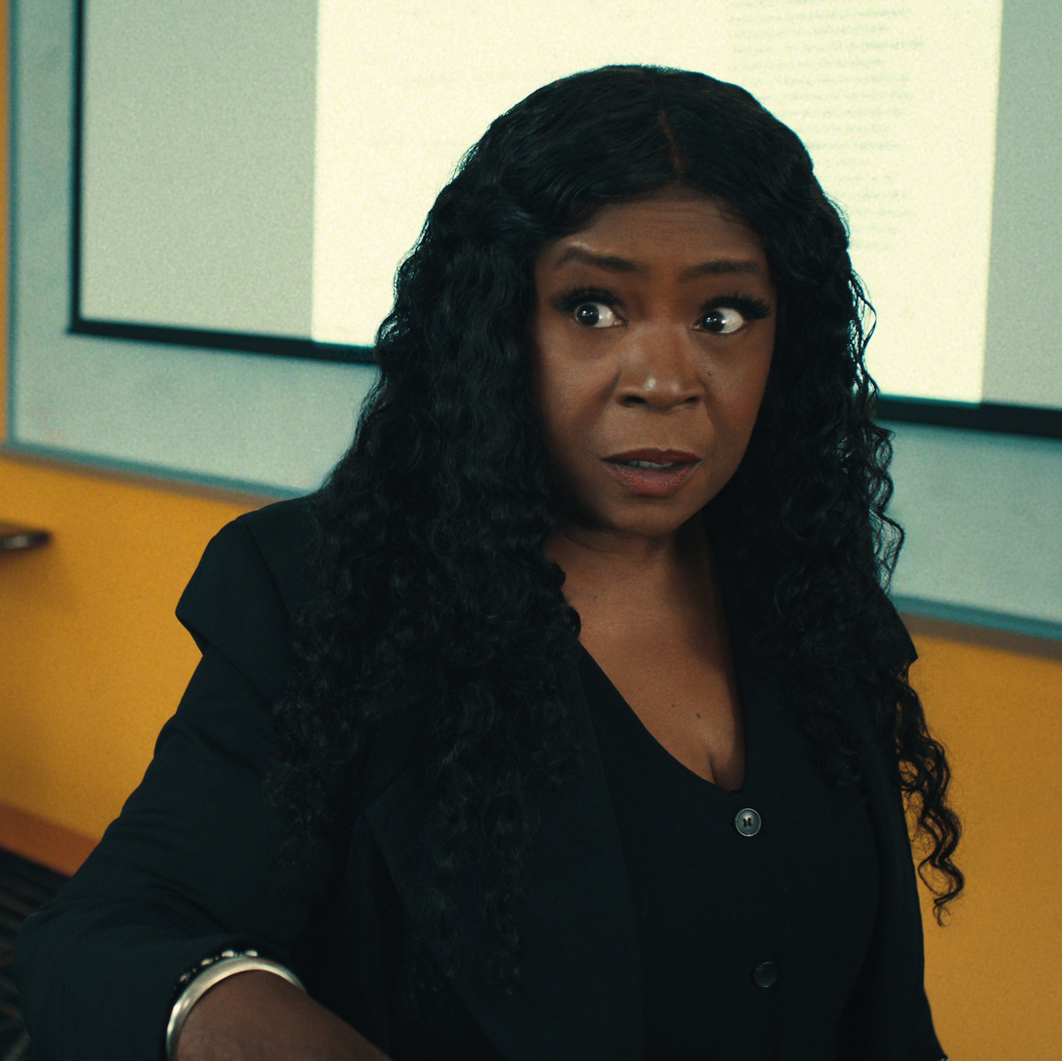 Woman with long curly hair wearing black blazer looks surprised in a classroom or office setting, with a projection screen and yellow wall in the background.