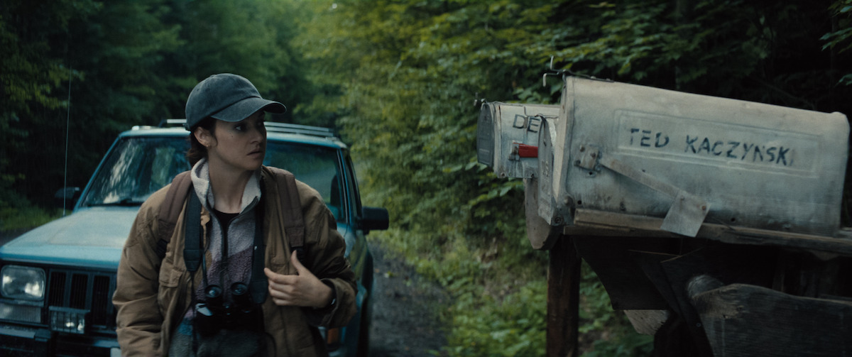 A person in outdoor clothing stands near a car on a forest path, looking at an old metal mailbox labeled "Ted Kaczynski," surrounded by dense green trees.