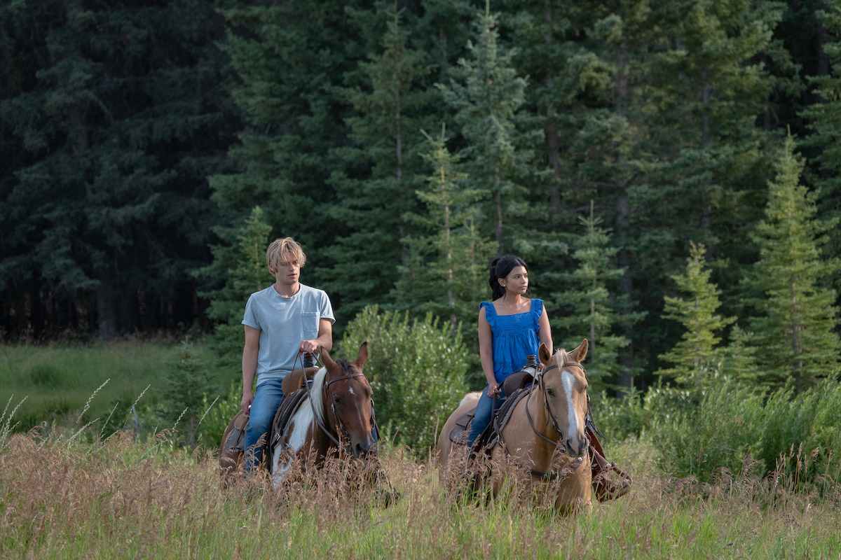Two people riding horses through a grassy field bordered by tall evergreen trees, both appearing relaxed and enjoying the natural outdoor setting in daylight.