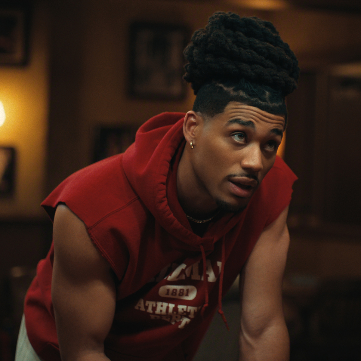 Athletic man with styled hair in a sleeveless red hoodie leans forward indoors, warm lighting, framed pictures in background, casual setting.