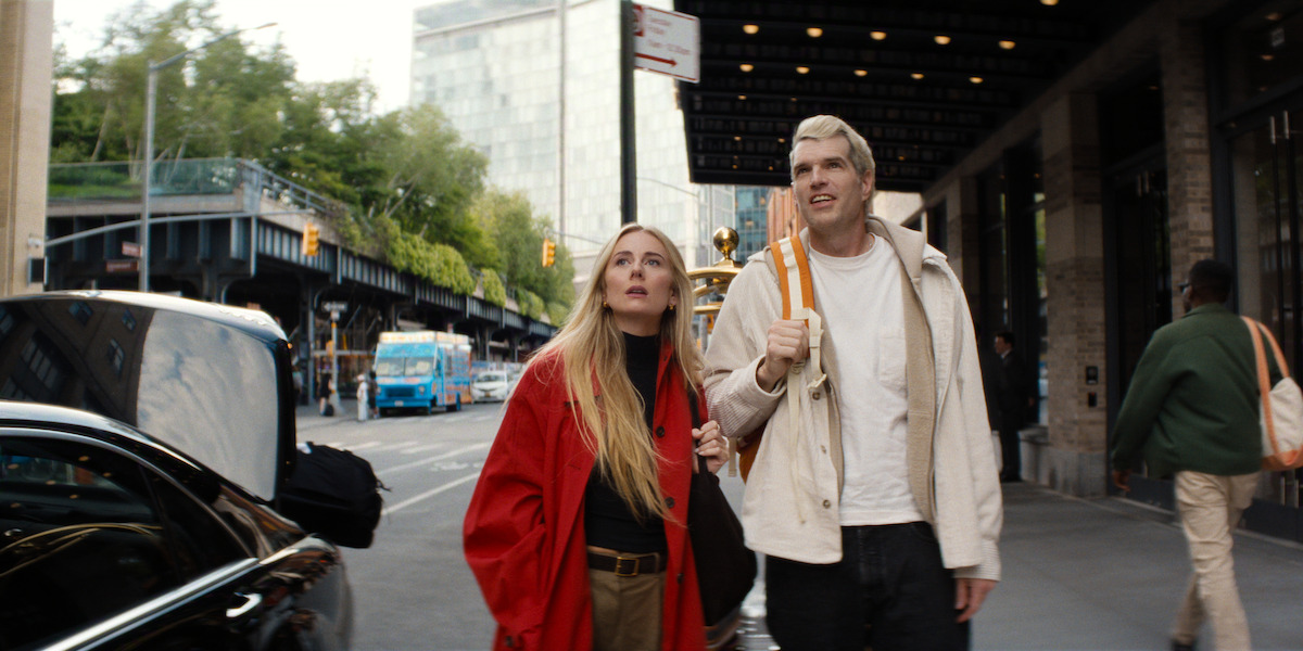 A man and woman walk side by side on a busy city sidewalk near a building entrance, both looking upward; trees, cars, and people are visible in the urban background.