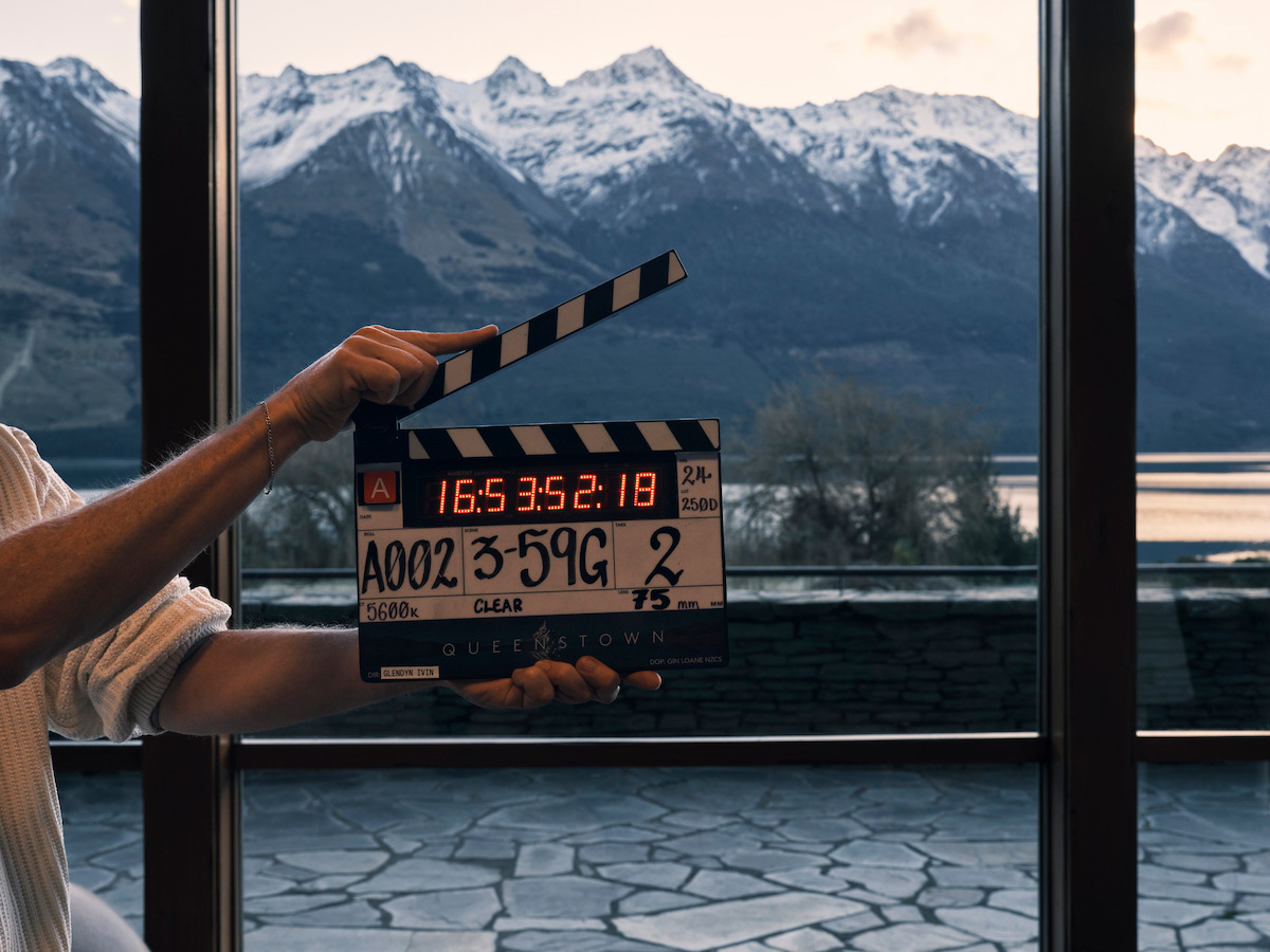A person holds a film clapperboard indoors in front of large windows, with a scenic view of a lake, stone terrace, and snow-capped mountains in the background during daylight.