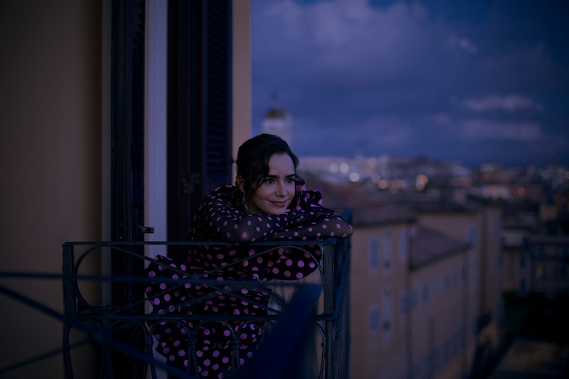 Lily Collins, como Emily, se apoia no guarda-corpo de uma sacada na temporada 4 de ‘Emily em Paris’