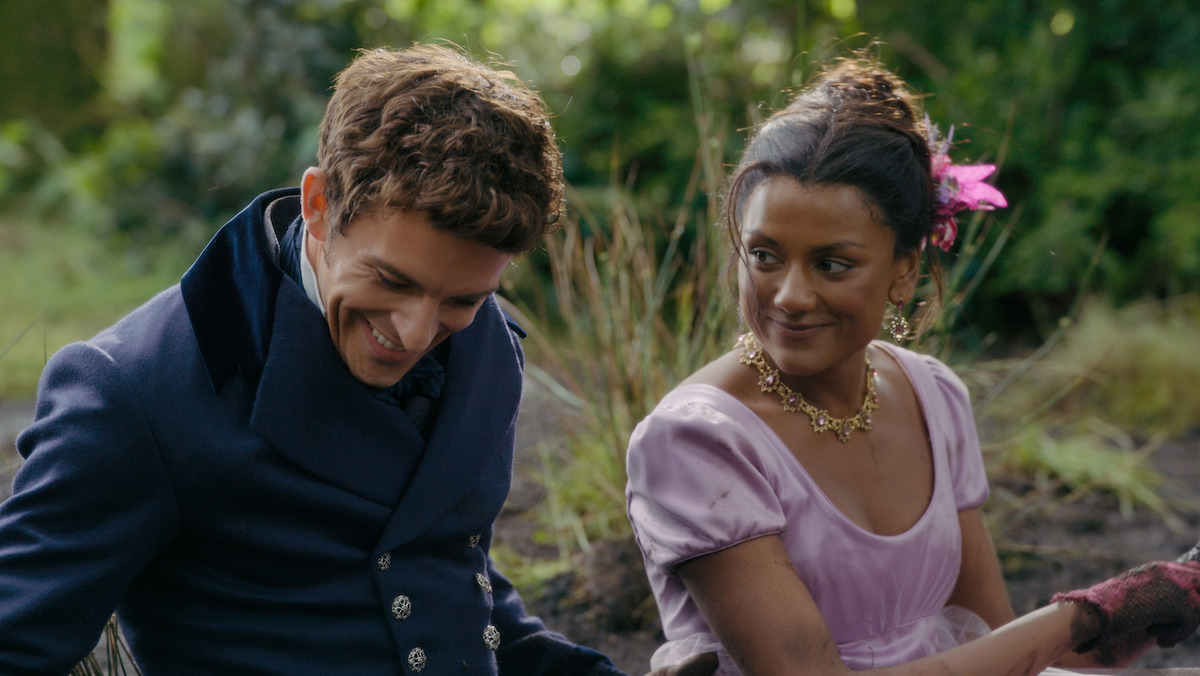 A smiling man in a dark blue Regency suit sits next to a woman in a light purple dress with a pink flower in her hair and gold necklace, outdoors in a green garden setting with sunlight filtering through trees.