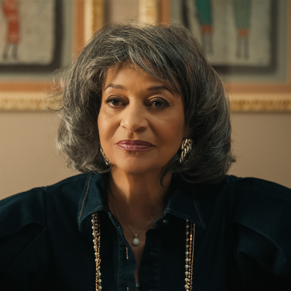 Older woman with gray hair wearing a dark shirt, gold earrings, and a necklace, seated indoors with framed art on a beige wall in the background. The lighting is warm and soft.