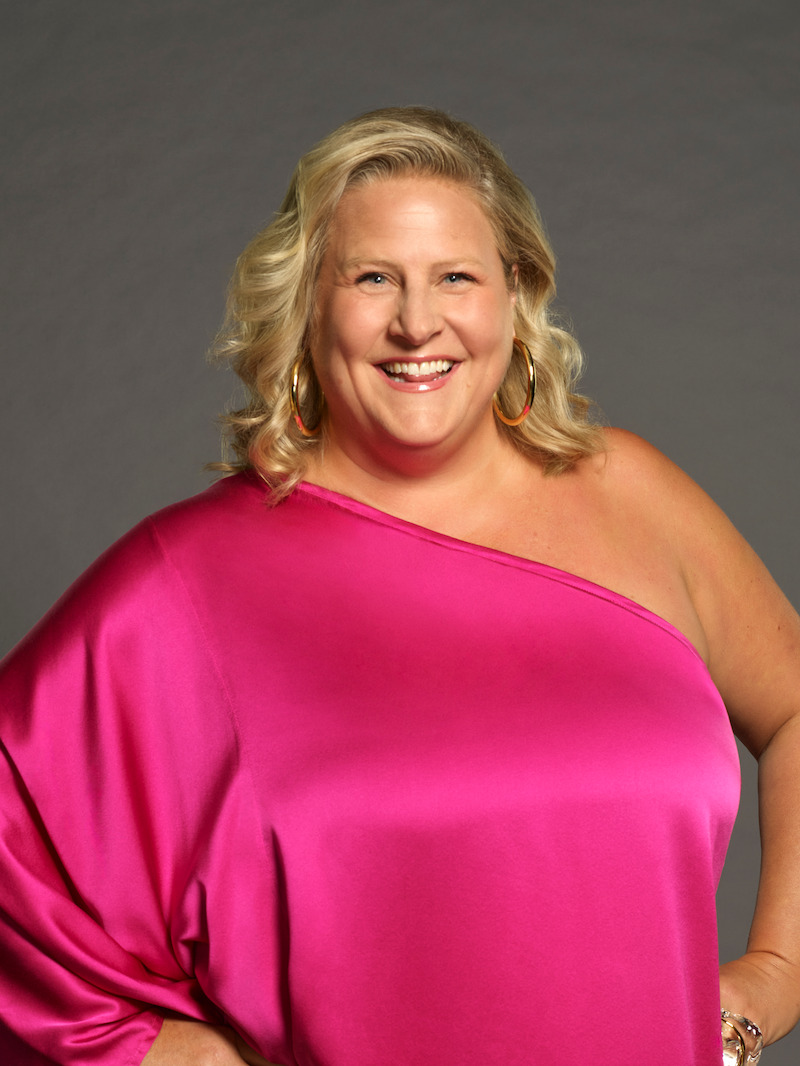 Woman with blonde hair wearing a bright pink one-shoulder dress, smiling and standing with hand on hip against a plain gray studio backdrop.