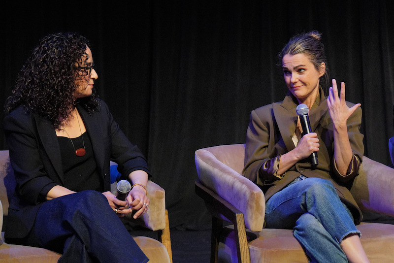 Two women sit in chairs on stage with microphones, engaged in conversation. The woman on the right gestures with her hand while speaking. They are in a dimly lit, indoor environment with a black curtain backdrop.