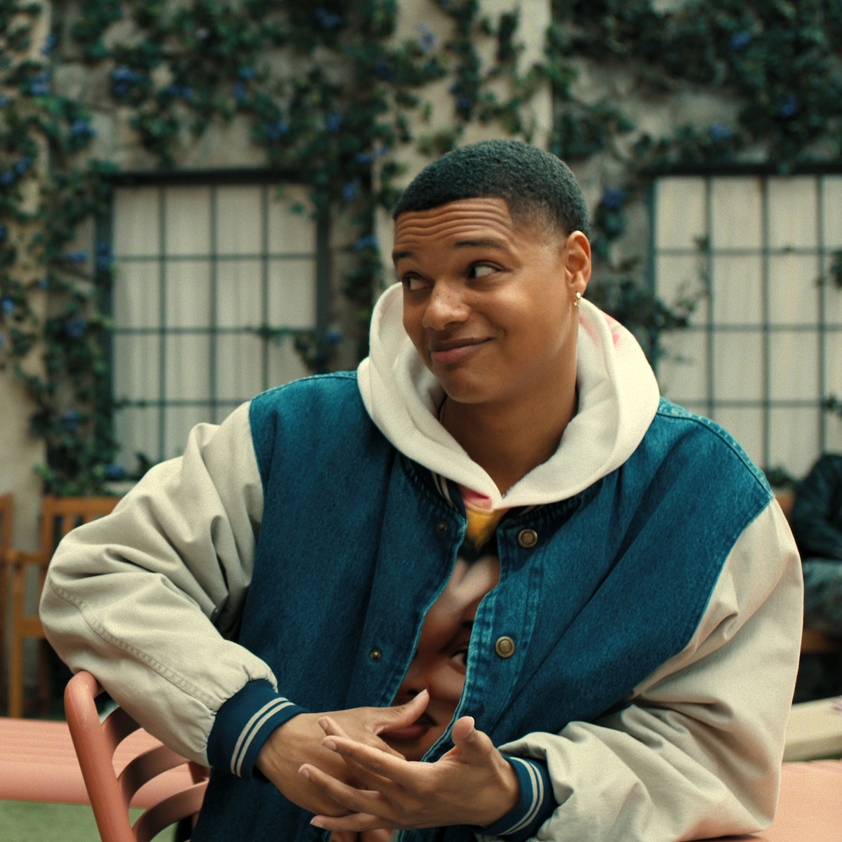 Person sitting outdoors on a pink chair, wearing a denim jacket over a hoodie, making a playful facial expression with hands gesturing. Background features a building wall with windows and green ivy or vine plants.