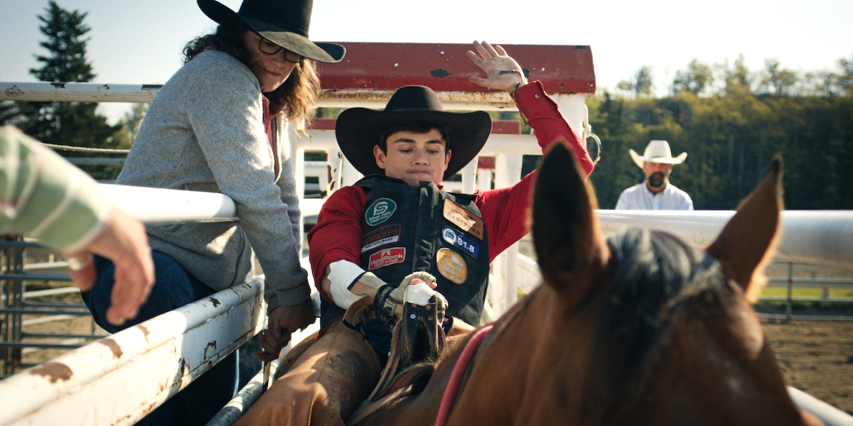 Young rodeo rider in full gear prepares to ride a horse in an outdoor rodeo arena, with a helper nearby and spectators in western hats in the background on a sunny day.