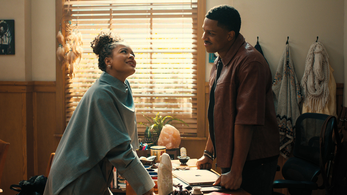 Two people smiling and talking in a cozy office with sunlight coming through blinds, desk with crystals and papers, wooden walls, plants, and hanging decor in the background.