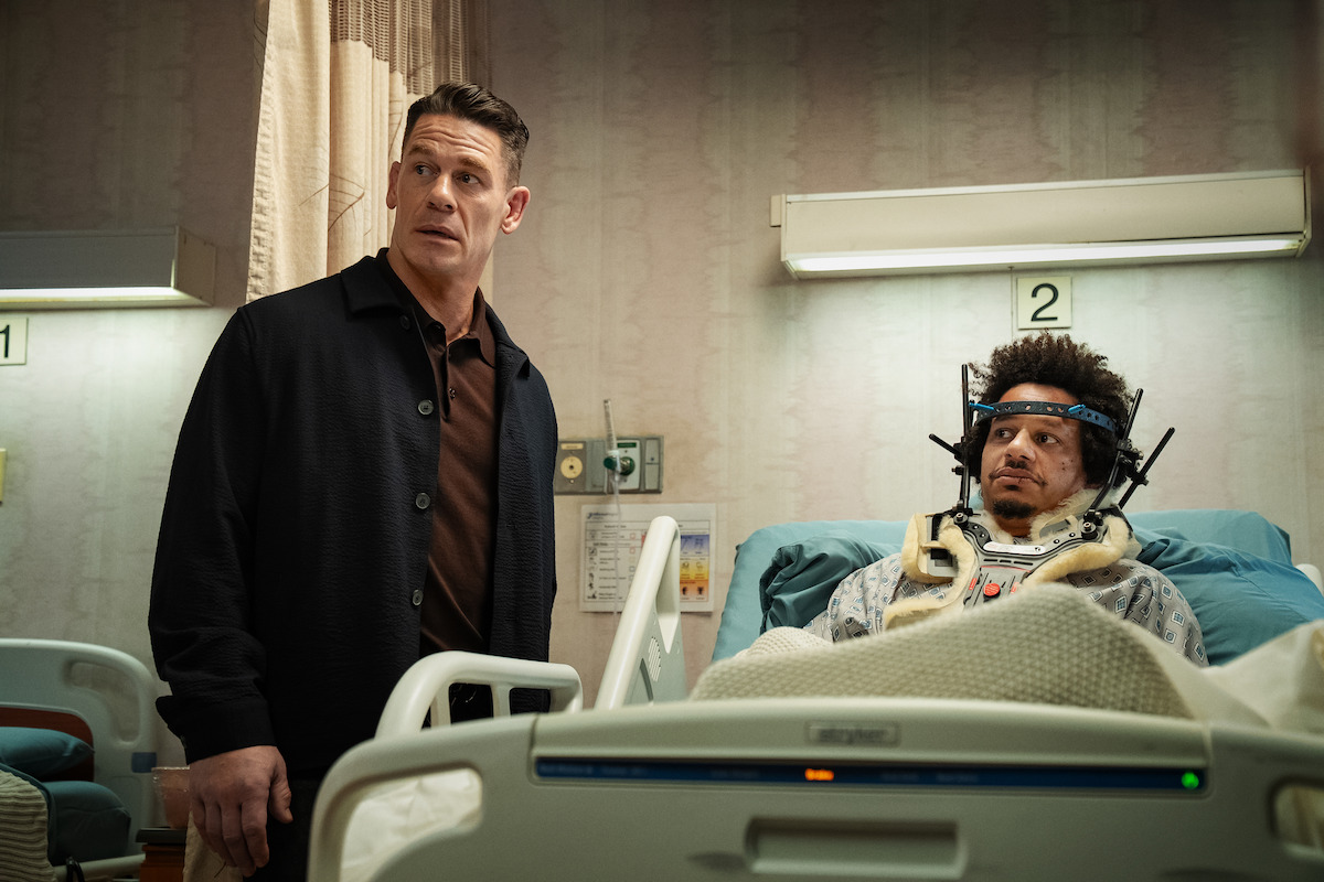 A man stands beside a hospital bed looking surprised while another person lies in bed with medical headgear, in a hospital room setting with numbered beds and medical equipment visible.