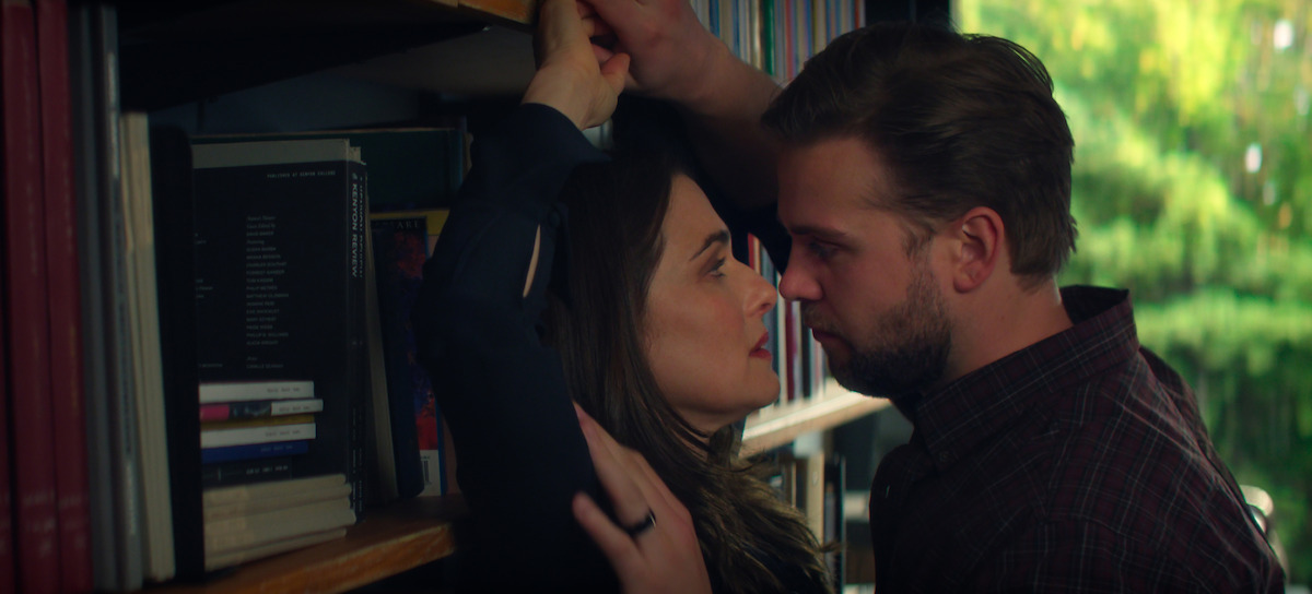 Rachel Weisz as M and Leo Woodall as Vladimir stand closely facing each other in a library, surrounded by shelves of books, with sunlight shining through a window in the background, creating an intense and dramatic atmosphere.