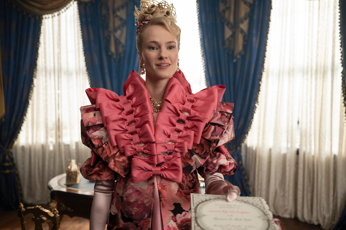 Uma mulher com um elaborado vestido rosa florido de estilo vitoriano está de pé e sorri enquanto segura um certificado. Ao redor, a sala luxuosa tem cortinas azuis e móveis de época.