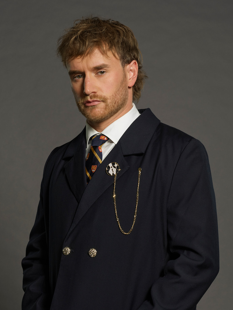 A man with light brown hair and a beard poses in front of a plain gray background, wearing a navy blue suit with a tie and gold chain accessory, giving a serious expression.
