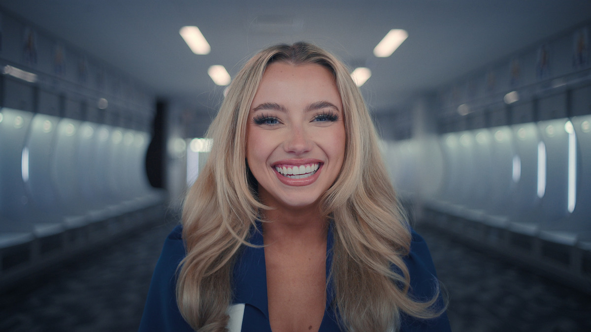 Confident woman smiles directly to camera and stands in an empty indoor sports stadium field, wearing a DCC uniform, with bright ceiling lights and empty seating in the background.