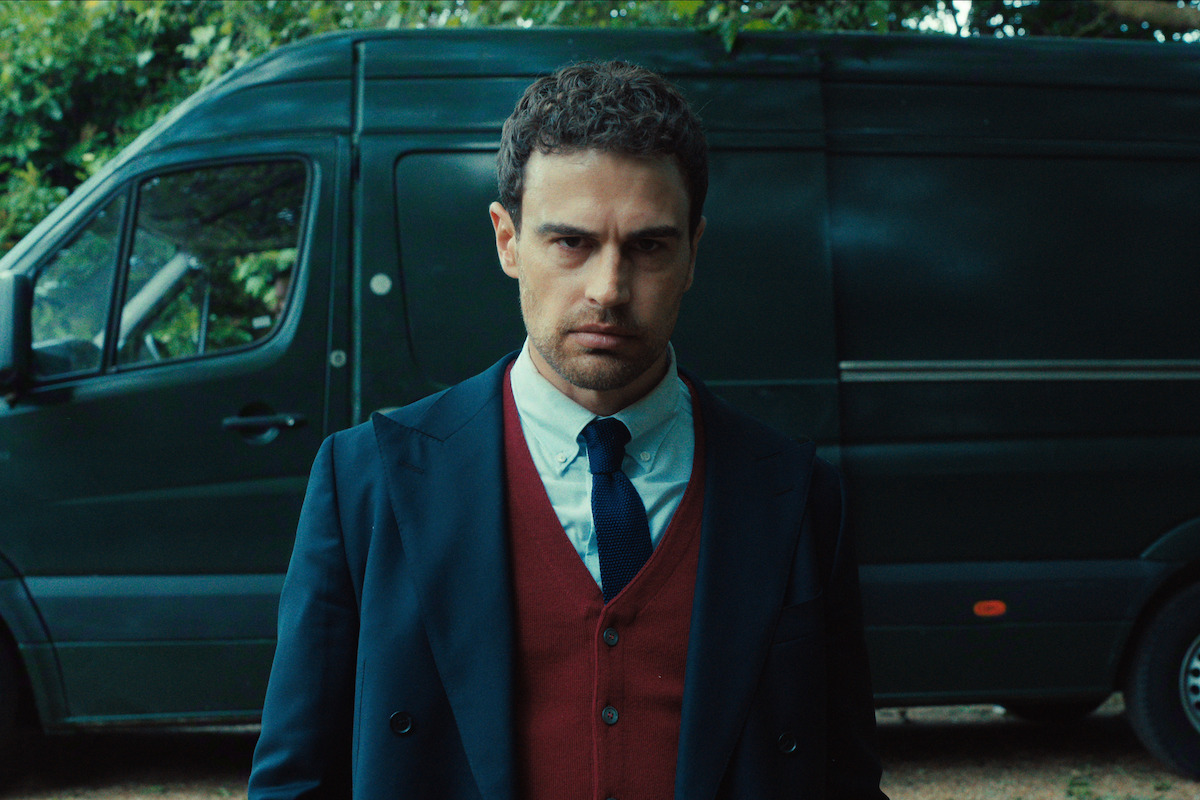 Serious man in a suit and red vest stands outdoors in front of a dark green van, with trees and greenery in the background, looking directly at the camera.