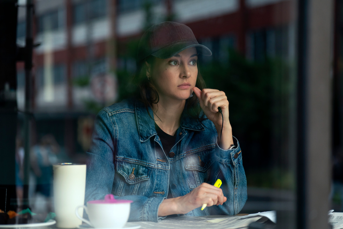 Woman in denim jacket and baseball cap sits at a table inside a cafe, looking thoughtful while holding a yellow highlighter, with a mug and papers in front of her, city street visible through the window.