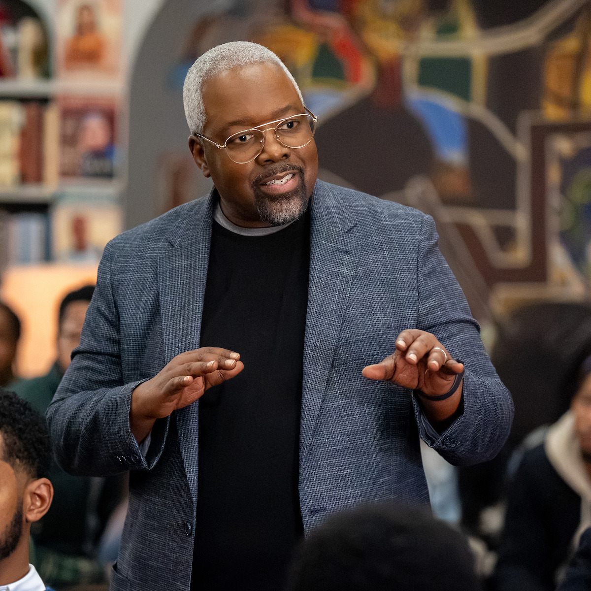 A middle-aged man with gray hair and glasses speaks to a group in a library or classroom, colorful mural and bookshelves in the background, suggesting an educational or discussion setting.