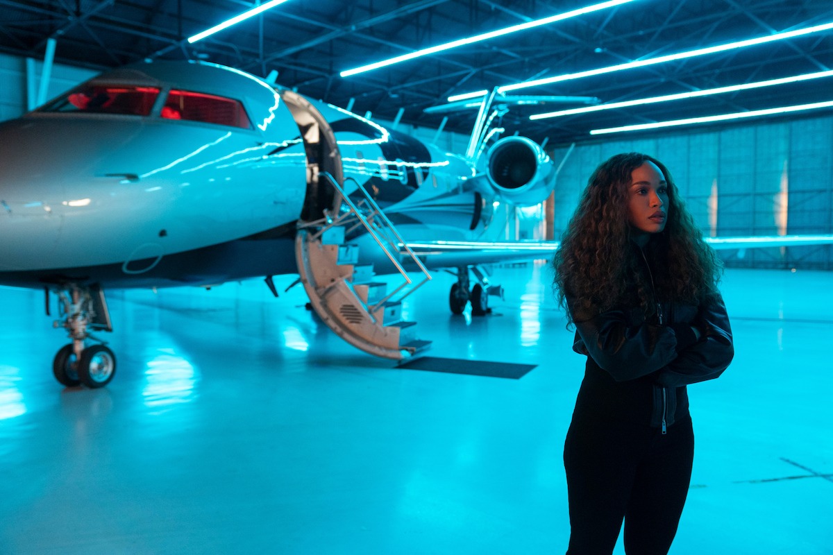Woman with curly hair stands confidently in a brightly lit, modern airplane hangar beside a sleek private jet, blue lighting reflecting off the glossy floor, giving a futuristic atmosphere.