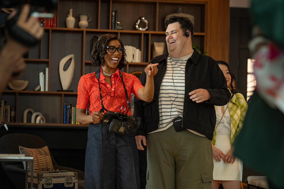 Two people laugh and interact on a modern set, with shelves and books in the background. The woman holds equipment, both wear headsets, suggesting a production or behind-the-scenes environment with a casual, friendly mood.