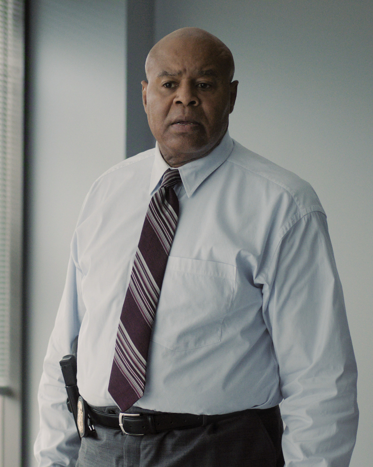 Man in a dress shirt and tie standing in a modern office with large windows and blinds, neutral expression, daylight coming through the windows.