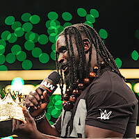 A wrestler with long dreadlocks and a beaded necklace speaks into a microphone labeled RAW, holding a golden crown. He wears a WWE t-shirt, standing in a brightly lit arena with green lights in the background.