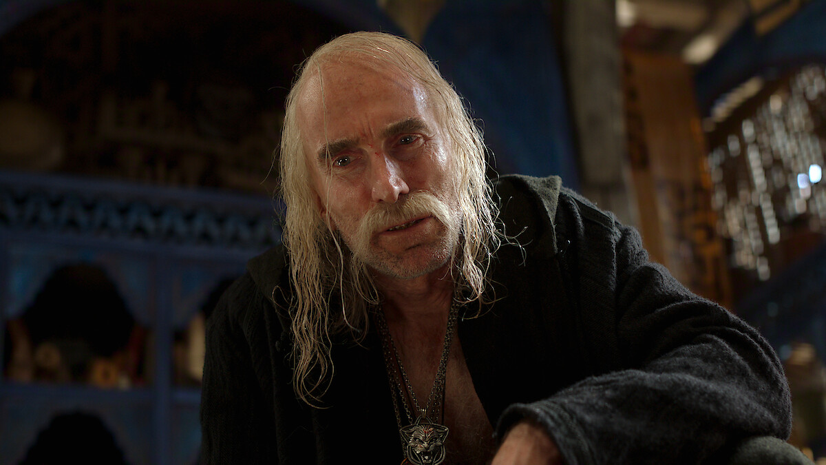 Elderly man with long white hair and mustache wearing dark robe, sitting indoors in a dimly lit room with blue and wooden details, serious and intense expression, medieval or fantasy setting, somber mood.