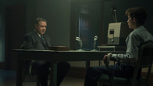 Two men sit across from each other in a dimly lit interrogation room, one appearing to question the other who is attached to a polygraph machine, with microphones and recording equipment on the table.
