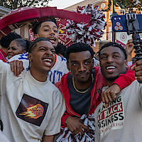 Three young men pose for a selfie at an outdoor event with a group, some wearing college-themed shirts, festive decorations and people with pom-poms in the background, creating a lively and celebratory atmosphere.