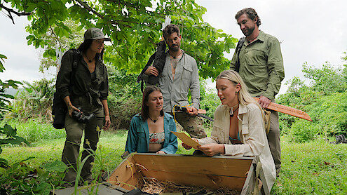 Five people outdoors in a green, forested area gathered around an open wooden chest; one person is holding papers and discussing findings as others watch attentively, with relaxed, adventurous expressions.