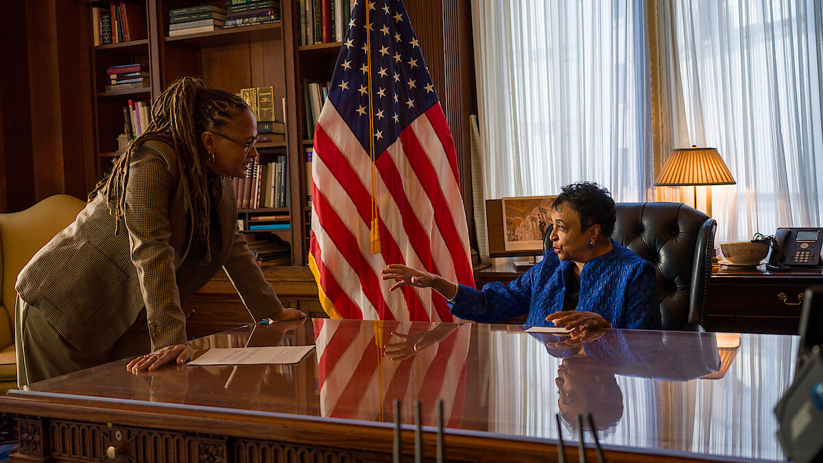 Two women in an office, one standing and one seated behind a polished desk, discuss something. An American flag, bookshelves, and a lamp are in the background, creating a formal, professional setting.