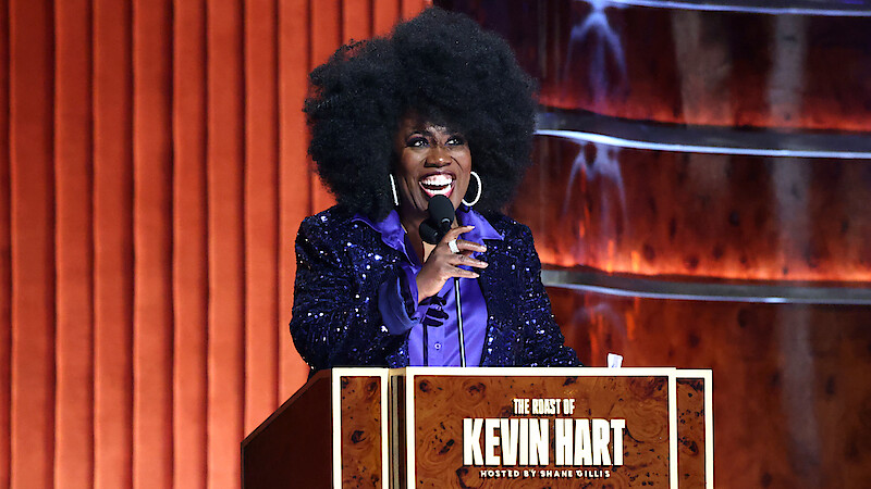  Sheryl Underwood with a large afro hairstyle and sparkly purple jacket speaks at a podium labeled "The Roast of Kevin Hart," on a stage with wood paneling and warm lighting.