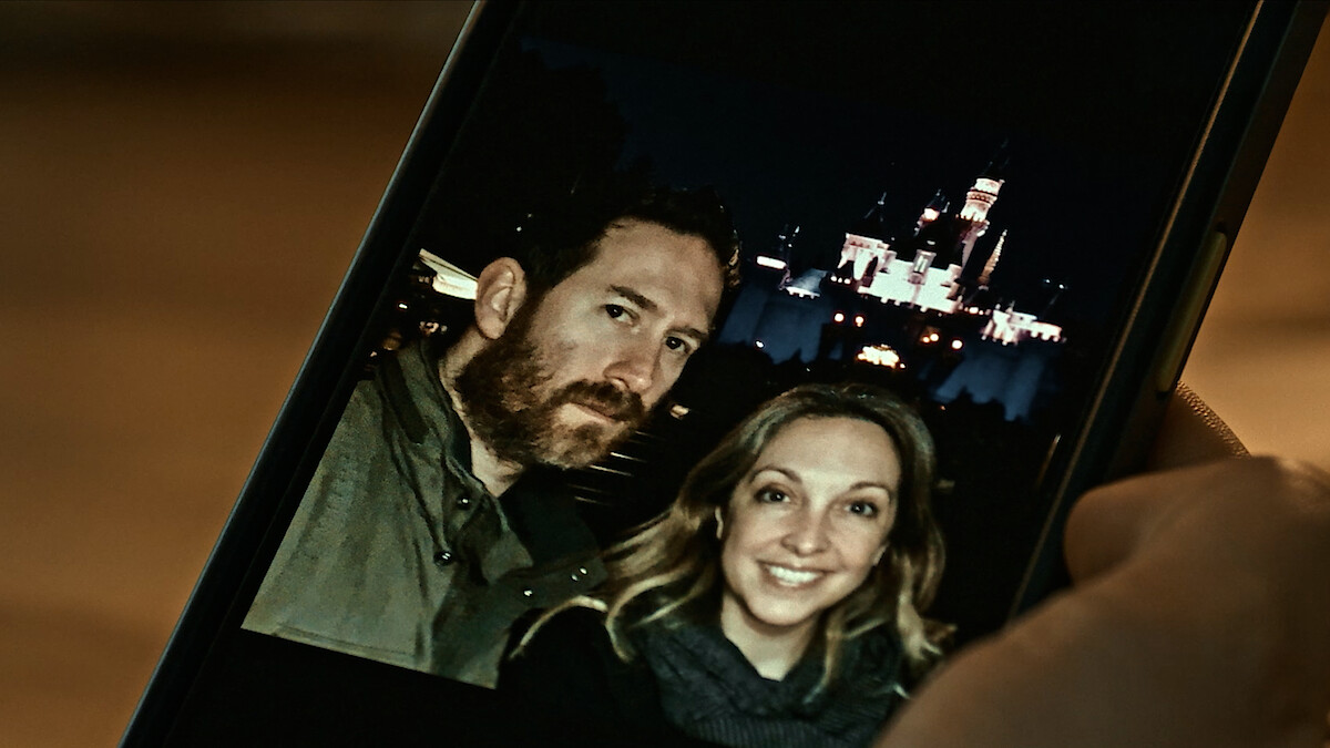 A man and woman smiling in a selfie photo on a smartphone, taken at night in front of an illuminated castle, likely at an amusement park. Hand holding phone visible in foreground.