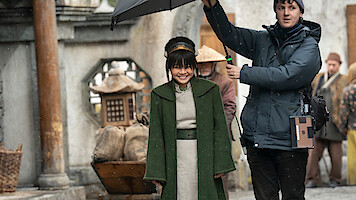 Miyako as Toph in a green coat stands outdoors on a film set while a crew member holds an umbrella over her, with people and rustic props in the background.