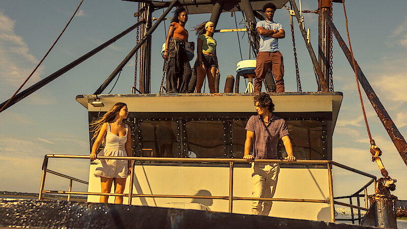 Five young adults standing on a boat under bright sunlight, with two people on the main deck and three on the upper deck, all enjoying a leisurely day on the water with a clear sky in the background.