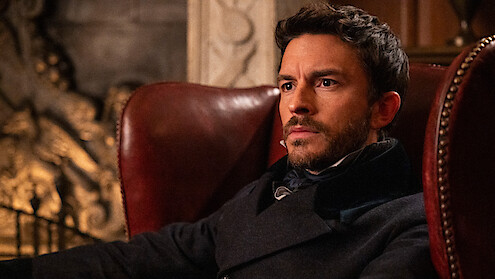 A man with a serious expression sits in a leather armchair in a richly decorated room with dim lighting and ornate details, suggesting a historical or formal setting.