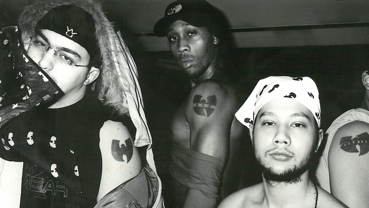 Três homens posam em uma foto em preto e branco em um ambiente interno, exibindo tatuagens do Wu-Tang Clan e roupas com o logotipo do grupo. O ambiente é descontraído, e eles usam streetwear urbano e bandanas, destacando sua ligação com o grupo e sua atitude.