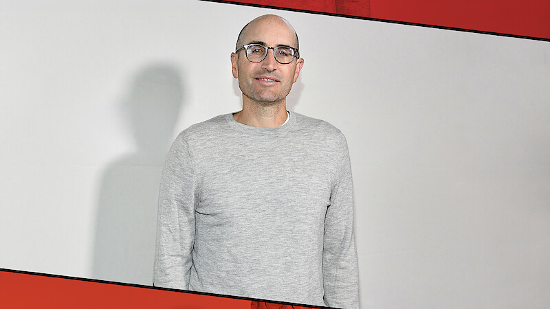 Man with glasses and a gray sweater stands in front of a plain white wall, casting a shadow, with a red and black diagonal border framing the image.