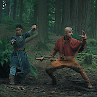 Two young people in fantasy warrior costumes pose in martial arts stances with focused expressions in a lush, green forest surrounded by tall trees and ferns, suggesting an adventurous or mystical setting.