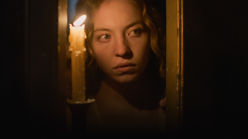 Woman with curly hair peering through a partially open door, illuminated by candlelight in a dark, dimly lit setting, creating a tense and mysterious atmosphere.