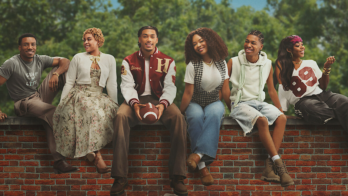 Six young adults sit on a brick wall outdoors, smiling and relaxed. Trees and blue sky in background. One holds a football; they wear casual, varied outfits, indicating a friendly, laid-back group setting.