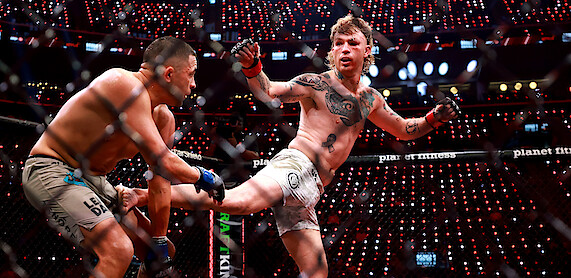 Two MMA fighters compete in a cage under bright arena lights; one delivers a high kick while the other defends, surrounded by a cheering audience and illuminated electronic displays.
